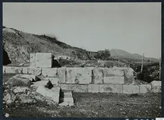 Large dressed limestone blocks of a wall and a stepped structure at Soli, with a hillside and wire fence behind.