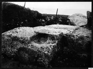 Large worn stone blocks at Soli, one with a rectangular hollow, with a grassy bank and wire fence behind