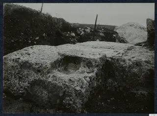 Large pitted stone block with a rectangular hollow in its top, at the theatre at Soli, with a fence post and stones behind.
