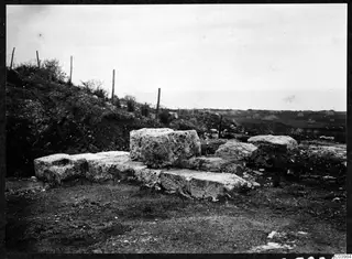 Pale cut stone blocks at Soli with a wire fence on the slope above and flat fields beyond.