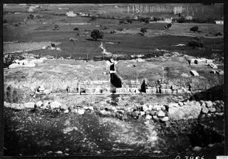 Excavated ruins of the theatre at Soli, with cut stone blocks and walls, a dry-stone wall in front, and fields and trees beyond