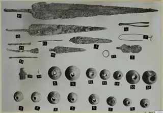 Metal blades, tools and a ring, with rows of perforated round objects, from Lapithos Tomb 322 B, each with a numbered label.