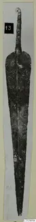 Corroded metal blade with a curled loop at the top, from Lapithos, photographed on a pale card strip.