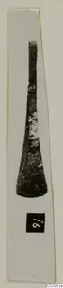 Bronze implement with a long narrow shank widening to a rounded blade, photographed upright on a white card strip