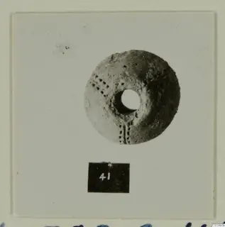 Pale disc-shaped find with a wide central hole and dotted impressions, photographed face-on on a mounted card with a number label.