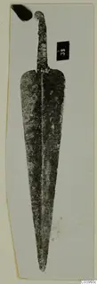 Corroded metal weapon blade from Lapithos, Vrysi tou Barba tombs, photographed on a pale card