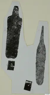 Two corroded bronze objects from Lapithos Tomb 313 C-D: a flat tool and a long pointed weapon.
