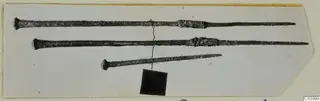 Three long metal pins or rods with knobbed heads and pointed tips, shown on a pale card mount.
