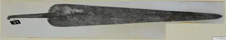 Long pointed metal blade with a slender tang at the left end, from Lapithos, shown on a card mount.