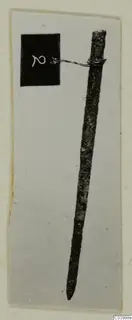 Long slender bronze metal tool tapering to a point, with a numbered tag wired to its blunt top end, from Lapithos Tomb 323 D.