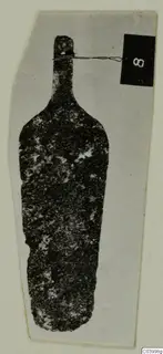 Corroded bronze tool from Lapithos Tomb 323 D, upright against a grey card, with a number label