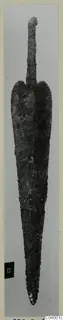 Corroded metal blade with a narrow tang, photographed upright against a grey background, from Lapithos