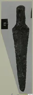 Corroded bronze blade-shaped tool from Lapithos, with a narrow tang, photographed on a grey card.