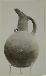 Pottery Red Polished III jug with a round body, tall neck, spout and loop handle, shown on a wire stand