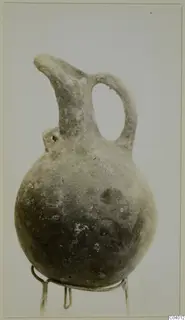 Red polished jug with a single loop handle and a pinched spout, shown on a wire stand against a plain background