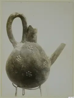 Red Polished III jug with a loop handle, broken neck and spout, standing on a wire stand