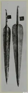 Two long metal blades with narrow tangs, photographed upright side by side against a grey background