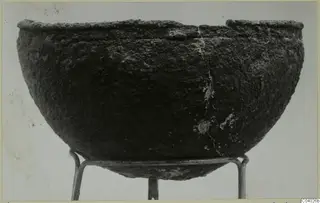 Deep rounded bowl with a dark, encrusted surface and broken rim, standing on a metal wire stand