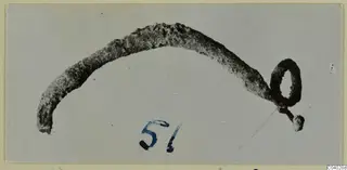 Corroded metal pin or rod with a loop at one end, photographed on a grey card with a blue number label.