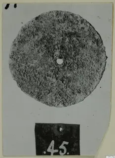 Corroded bronze disc with a central hole, photographed on a card with a number label below it.