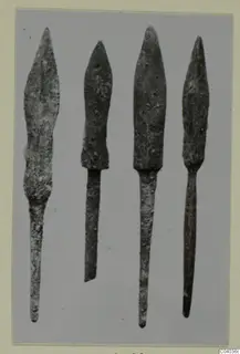 Four pointed metal blades with long tapering tangs, standing side by side against a grey background.