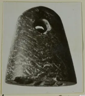 Dark stone object with a round perforation near the top, photographed on a pale card mount