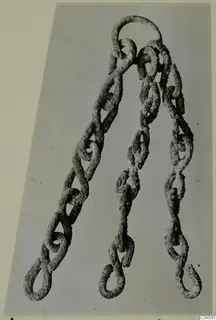 Corroded metal chain with a ring at the top and three branches ending in loops, laid flat on a grey backing
