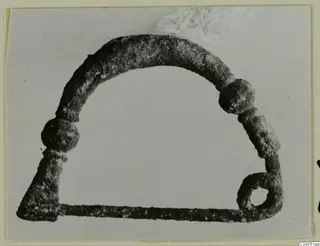 Corroded metal loop with two knobs on the arch and a ring with a hole at one end, photographed on a pale background.