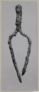 Corroded metal fragment from Idalion, a stem splitting into two long arms, shown flat against a pale background