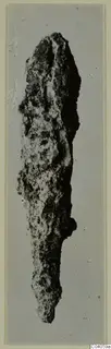 Heavily corroded iron object, long and narrow, photographed upright against a plain grey backdrop