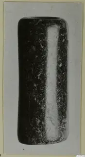 Dark stone object shaped like a short cylinder with a pale vertical band, shown on a grey card mount