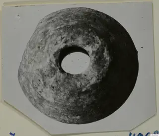 Disc-shaped stone object with a large central hole, shown in a photograph mounted on a card