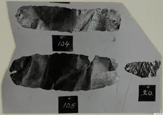 Three thin, crumpled metal sheet fragments from Enkomi Tomb 6, each with a small hole, laid on grey card