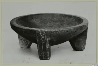 Stone bowl standing on three short legs, one leg with a band of circular motifs carved down it.