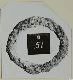 Corroded ring-shaped metal object on a white card, with a black number label placed in its centre