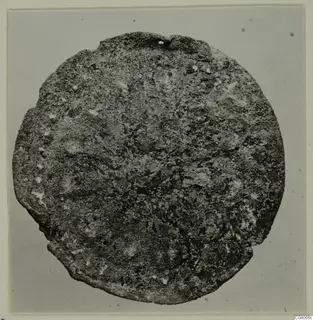 Corroded round metal disc with a small hole at its upper edge, photographed face-on on a grey background.