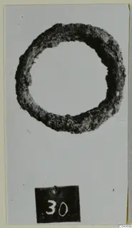 Corroded ring-shaped metal object from Enkomi Tomb 18, photographed on a light card with a number label.