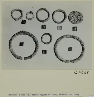 Enkomi Tomb 18: corroded metal rings and a pitted disc-shaped lump, each beside a number label, laid out on a card.