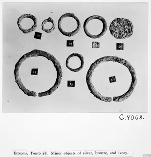 Eight corroded rings and one lumpy disc-shaped object from Enkomi Tomb 18, each set beside a numbered label