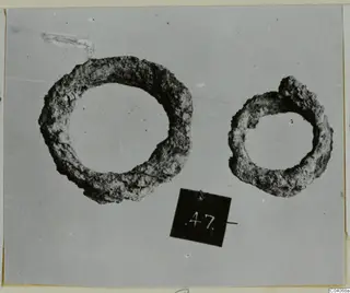 Two corroded ring-shaped metal objects with a number label, photographed from above, from Enkomi Tomb 18