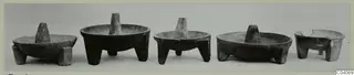 Five dark stone bowls on short legs, each with a central upright pestle, set in a row against a plain background