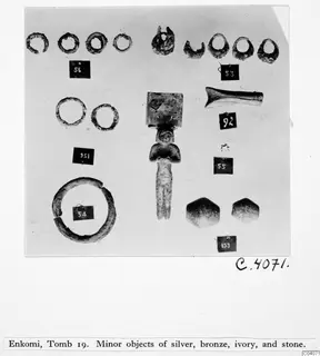 Small finds from Enkomi tomb 19: rings, a small standing figurine with a square plate, a curved pale piece and two stone objects.