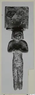 Small human figurine with folded arms and a flat plate with holes on top, photographed on a pale card mount