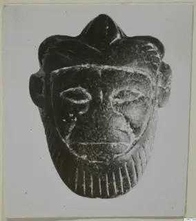 Dark stone face-shaped object with a pointed crest, projecting ears and almond-shaped eyes.