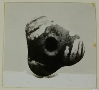 Dark perforated object, possibly a bead, with pale banded markings, photographed against a light background