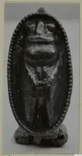 Small oval pendant with a beaded rim and a face-like relief, shown against a grey backdrop.