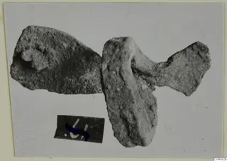 Two corroded bronze mounting fragments on a plain grey background, with a small number label below