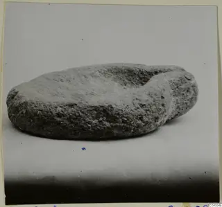 Rough stone object with a shallow hollow on top, photographed on a pale surface.