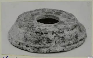 Squat grey object with a large round central hole and a ridged, stepped edge, photographed on a pale background