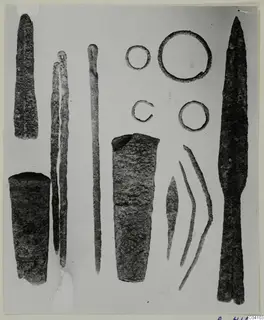 Metal weapons and tools from Arsos: a spearhead, two tapering tubes, long rods, rings and a small blade, laid out on a pale surface.